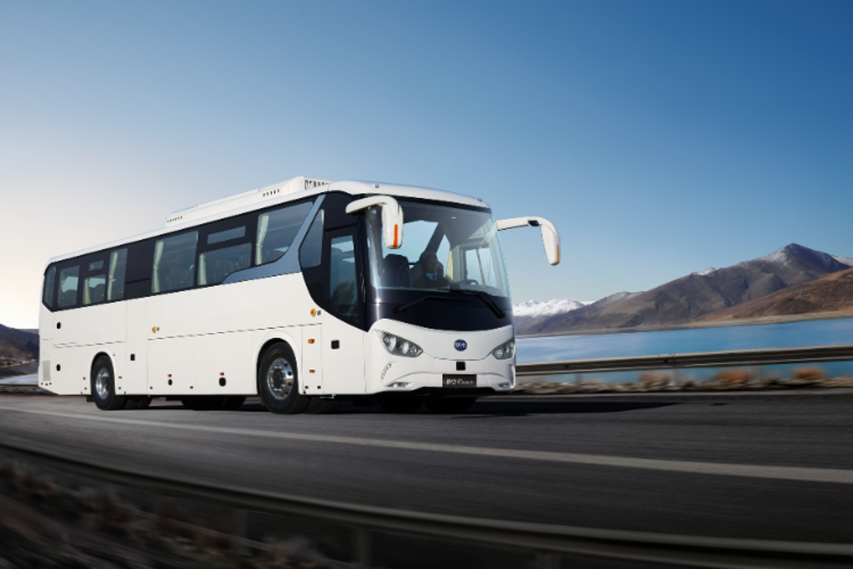 Ein weißer E-Reisebus fährt auf einer Straße. Im Hintergrund sind ein See und Berge zu sehen.