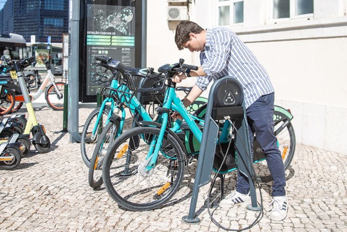 A man locks a bicycle to use it. Buses can be seen in the background.