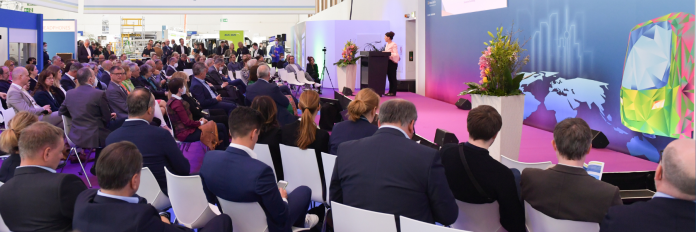 Auf einer Bühne findet eine Podiumsdiskussion mit sechs Teilnehmer:innen statt. Im Vordergrund ist das Publikum zu sehen.