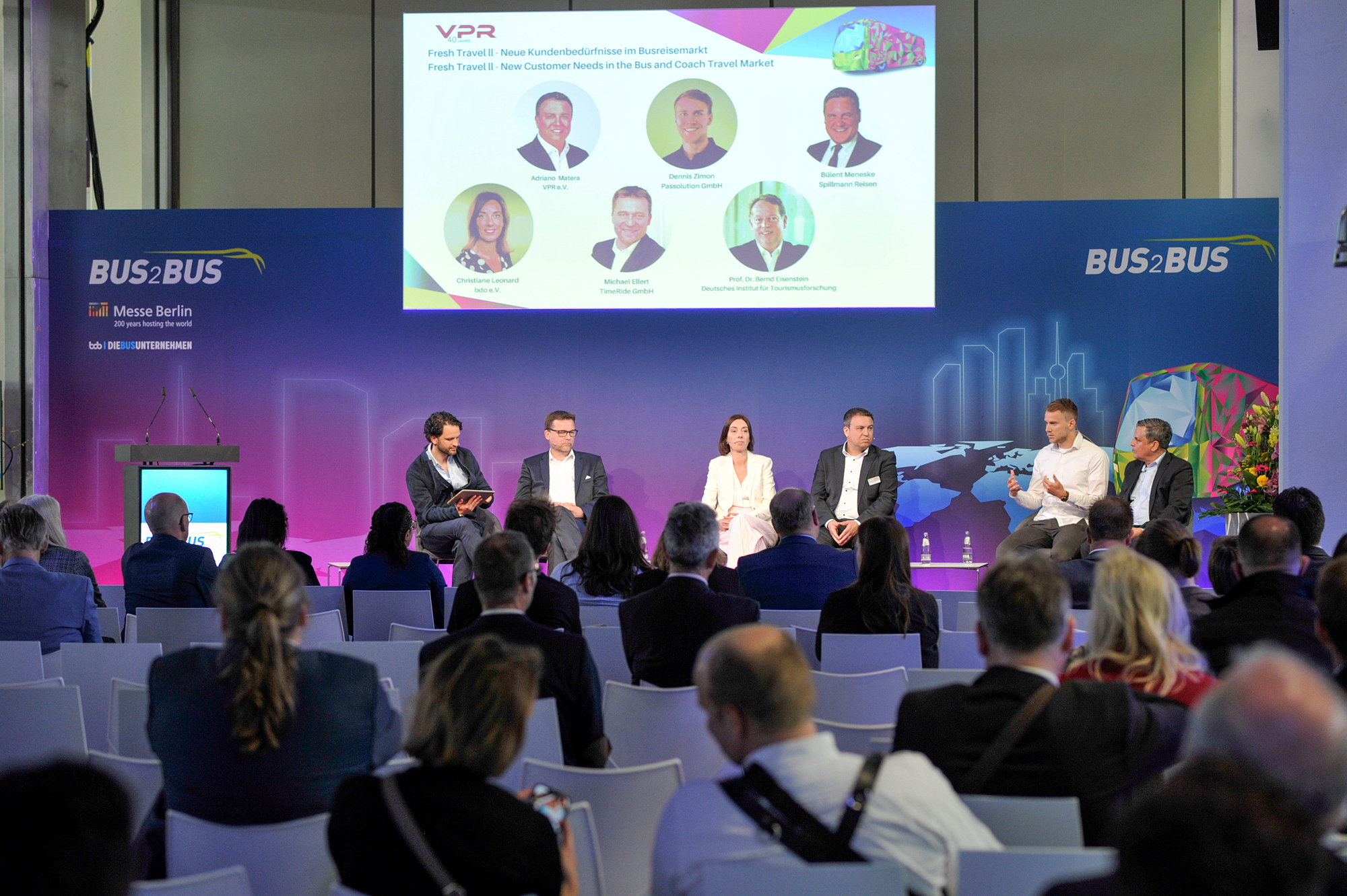 A panel discussion with six panellists takes place onstage. The audience can be seen in the foreground. 